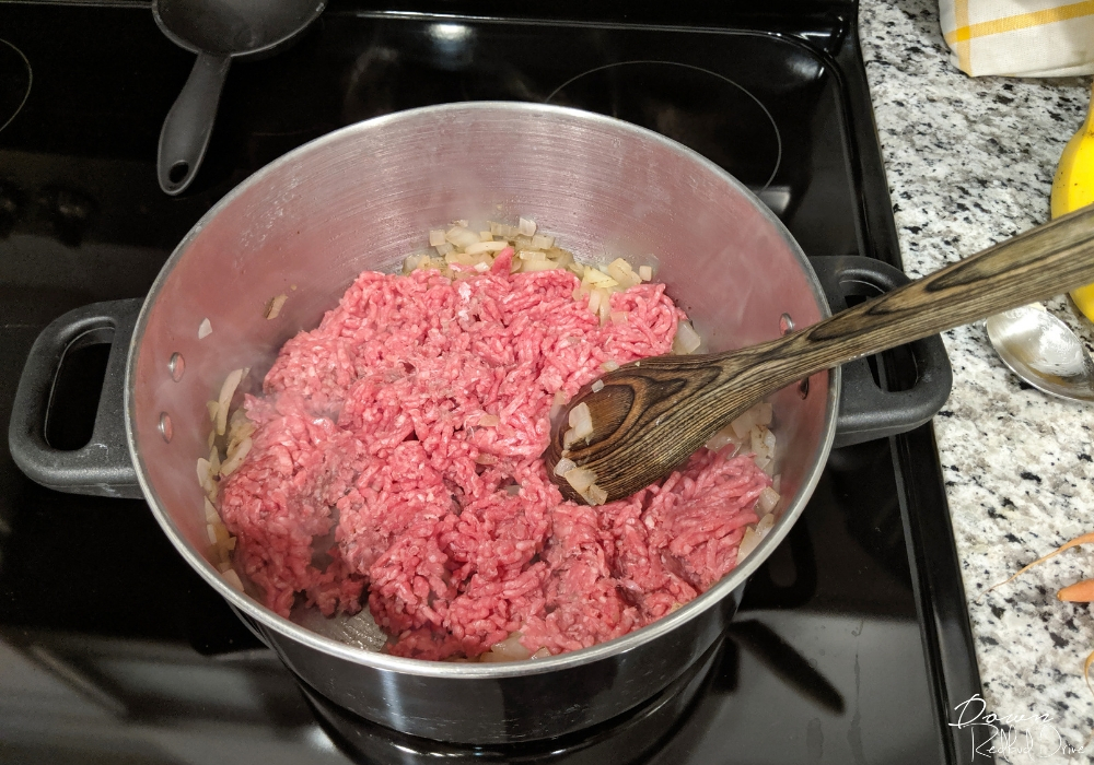 raw hamburger in a pot on the stove with chopped onion and a wooden spoon