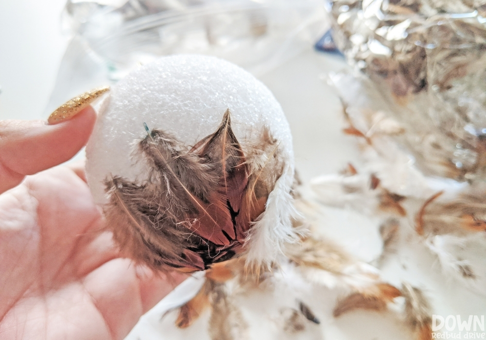 Up close picture of feathers being attached to a foam ball for the feather ornament DIY.