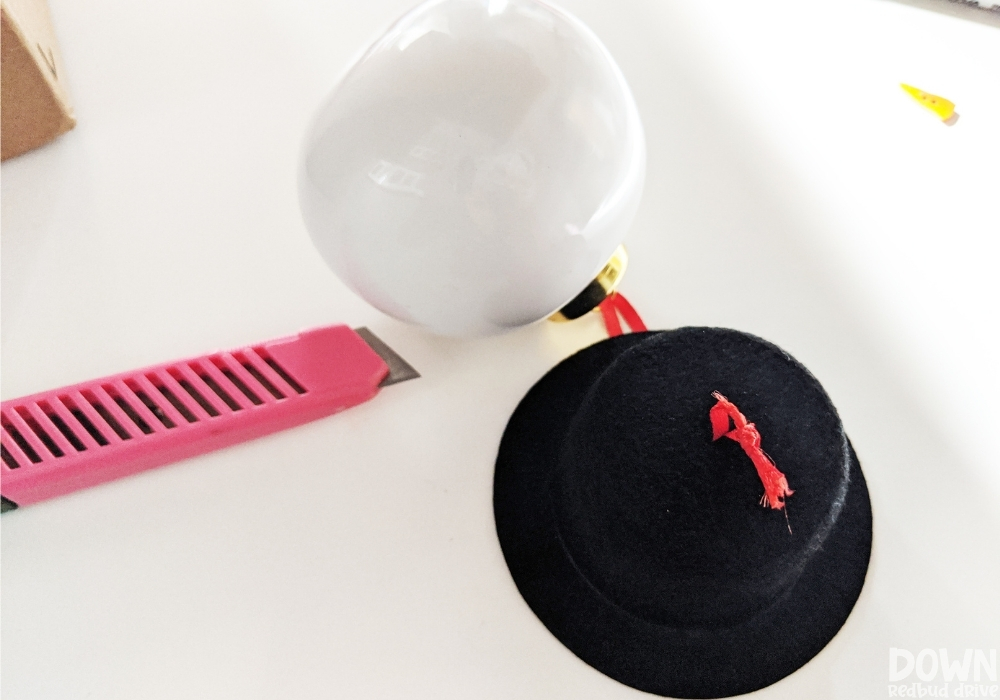 The red ribbon from a white Christmas ornament being strung through a hole in a small felt top hat.