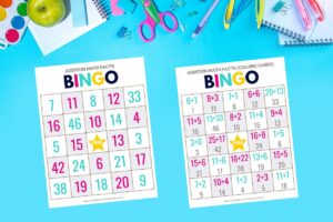 2 Printed bingo sheets from the addition math facts bingo game for kids free download on a bright blue background with scissors, pins, and other school supplies surrounding the page