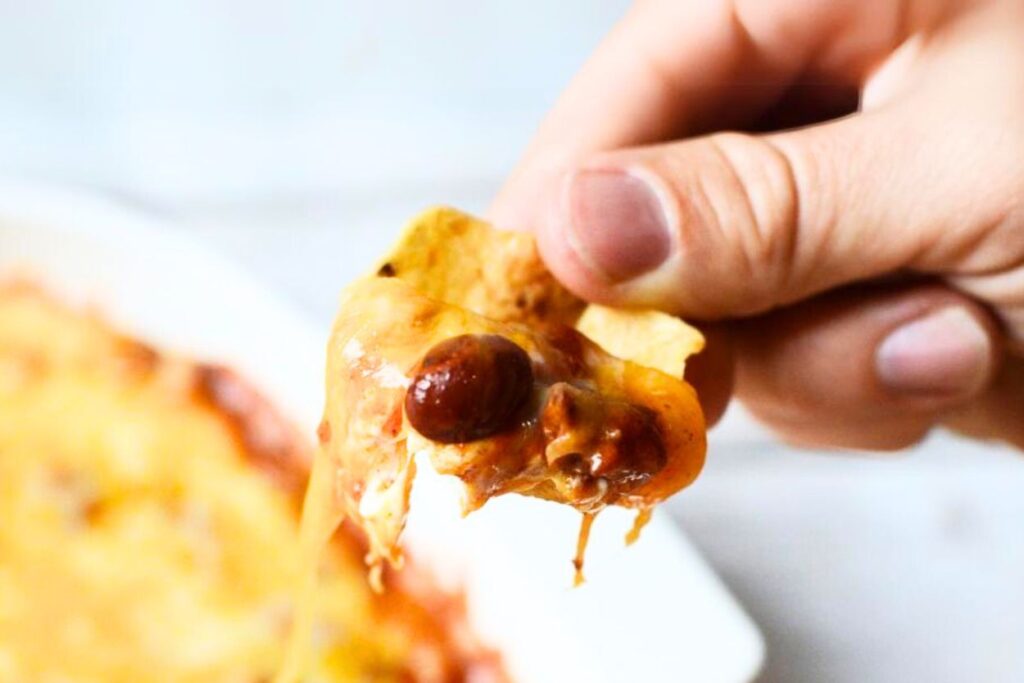 a corn chip coated in chili cheese dip being held above the finished pan of baked chili cheese dip