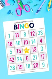 Printed bingo sheet from the addition math facts bingo game for kids free download on a bright blue background with scissors, pins, and other school supplies surrounding the page