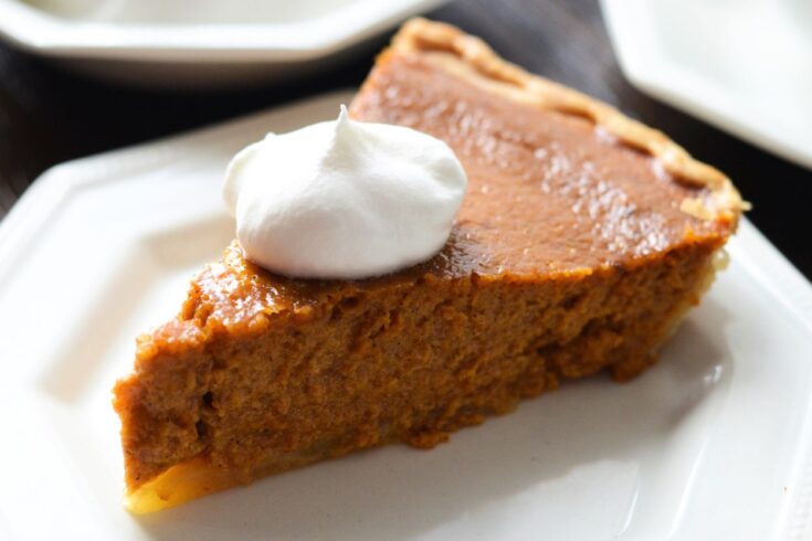 Slice of homemade pumpkin pie on a white plate, a cup of coffee and the remaining pie in the background
